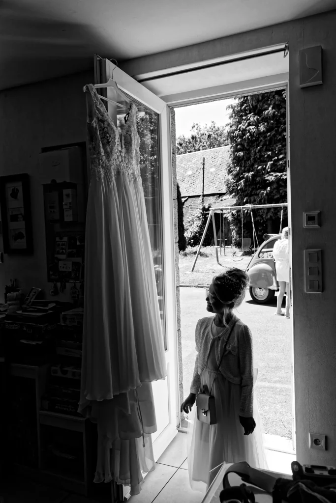 Enfant d'honneur en noir et blanc admirant une robe de mariée suspendue à Saint-Hilaire-du-Harcouët, Normandie.
