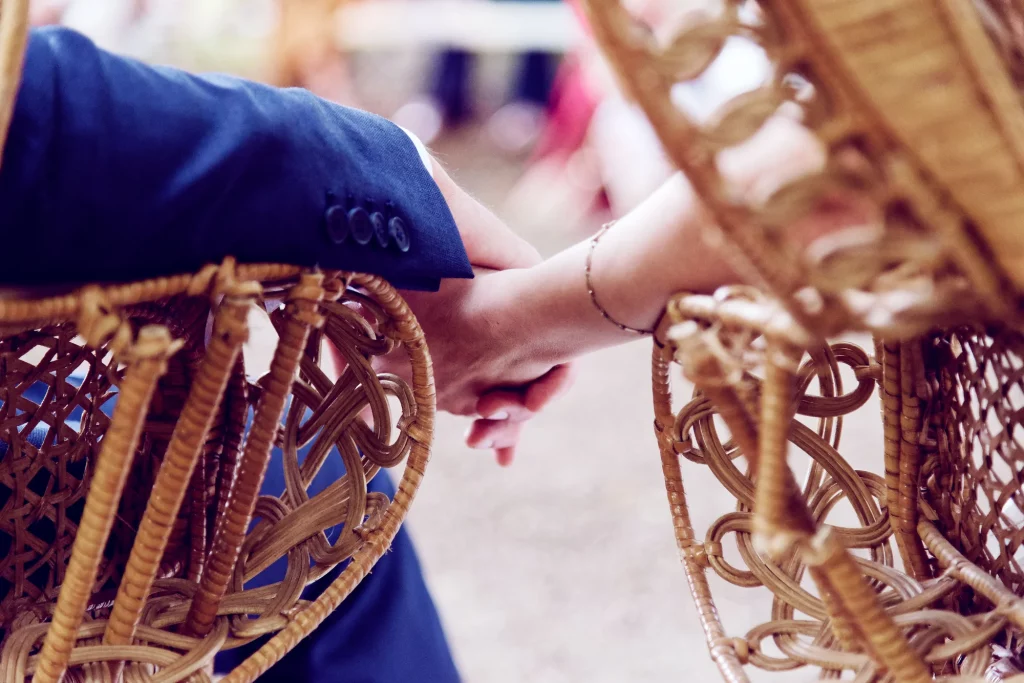 Gros plan de mains jointes dans des fauteuils en osier, mariage en Normandie à Saint-Hilaire-du-Harcouët.