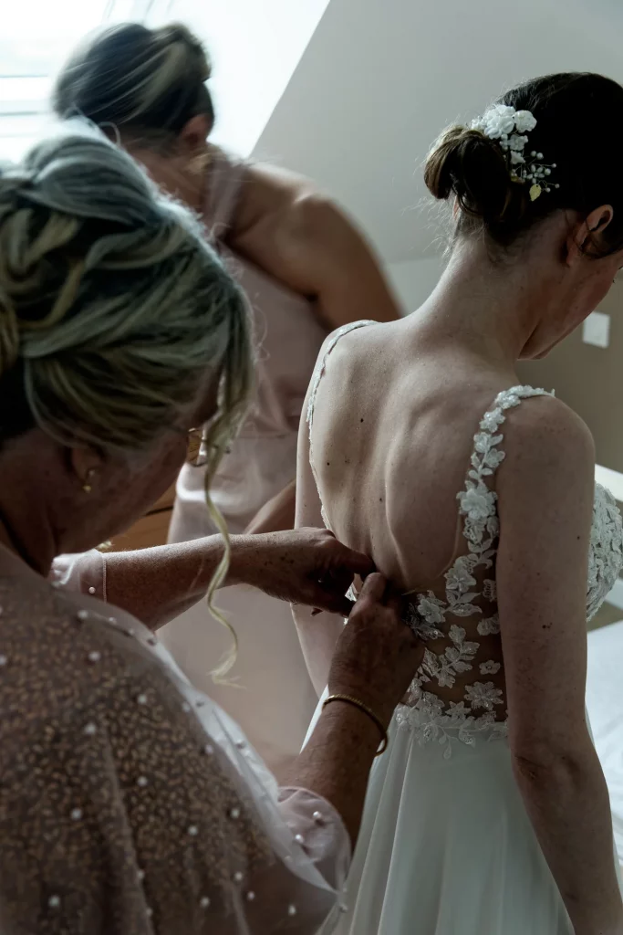 Gros plan sur une mariée en Normandie dont on boutonne la robe de dentelle dos nu pendant les préparatifs.