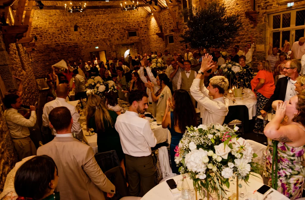 Invités debout applaudissant pendant un dîner de mariage en Normandie dans une grange en pierres.