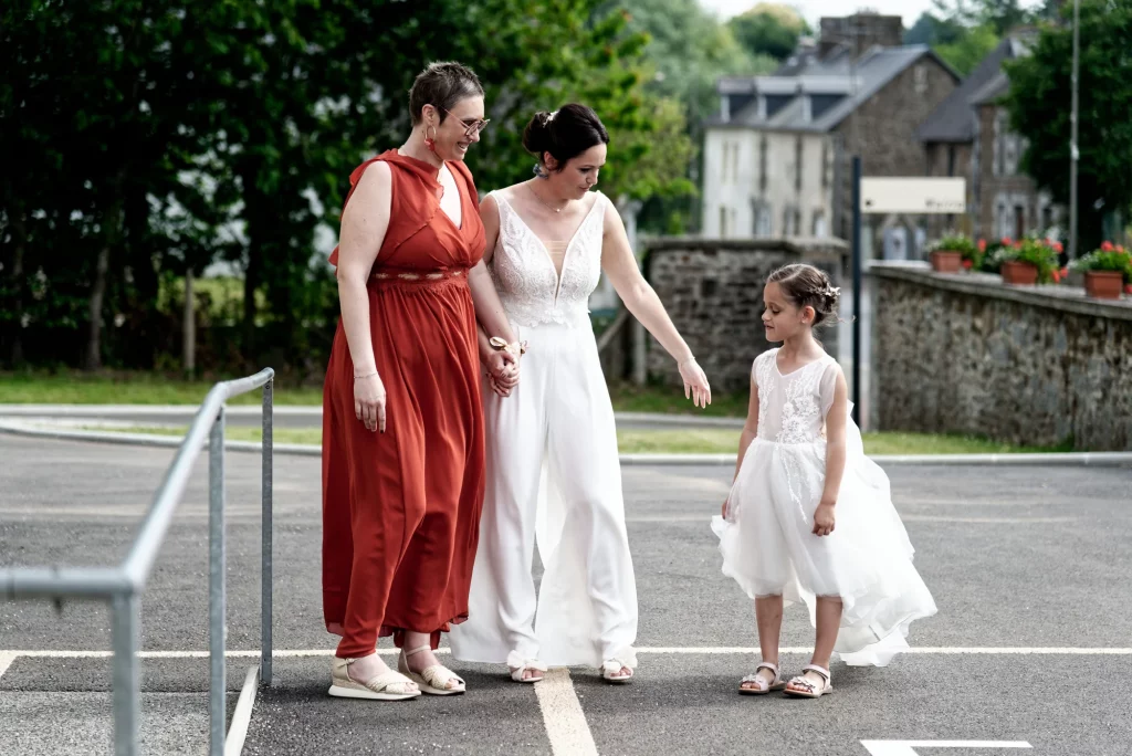 La mariée en blanc et une amie en robe rouille regardent une fillette à Saint-Hilaire-du-Harcouët.