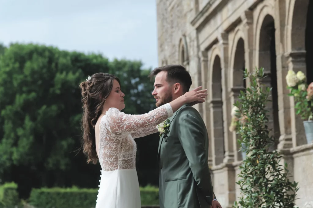 Mariée en dentelle ajuste le costume vert du marié devant un manoir en Normandie.