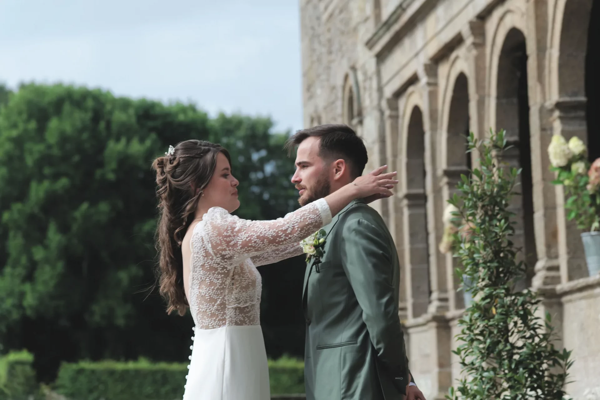 Mariée en dentelle ajuste le costume vert du marié devant un manoir en Normandie.