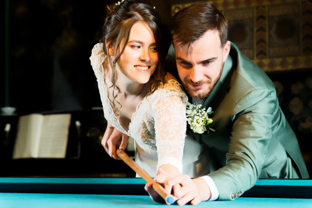 Mariée en dentelle et marié en costume vert jouant au billard à Saint-Hilaire-du-Harcouët.