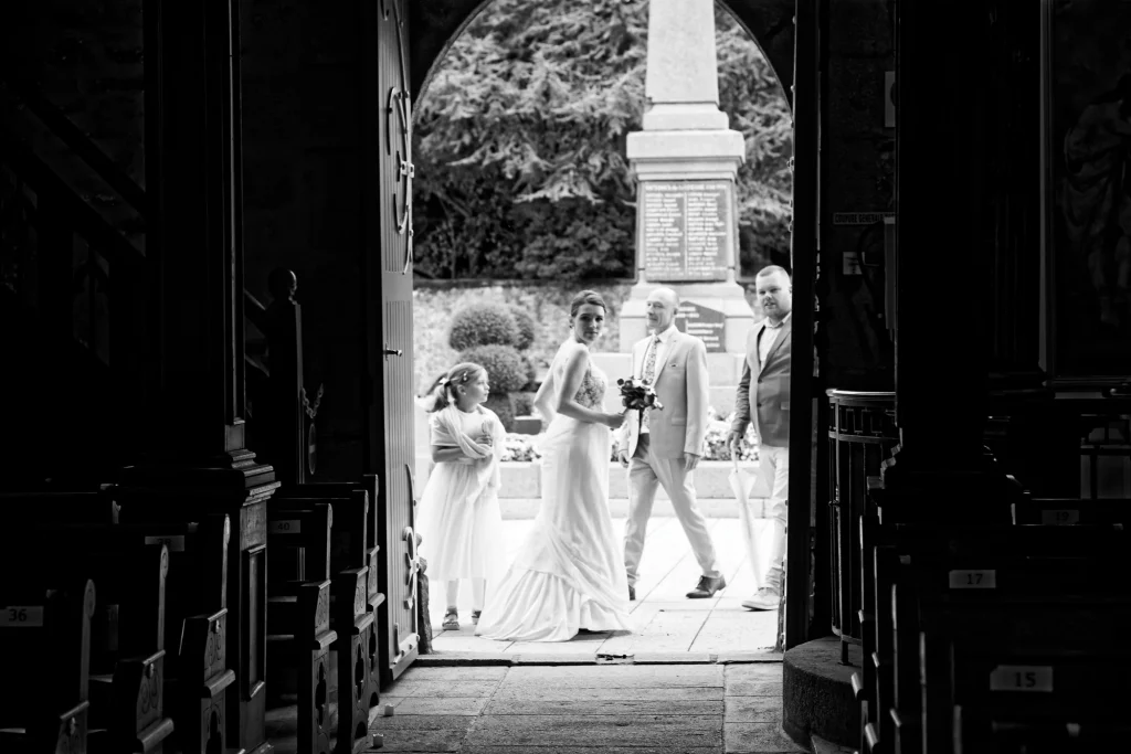 Mariée en robe blanche regarde en arrière à l'entrée d'une église en Normandie, style noir et blanc.