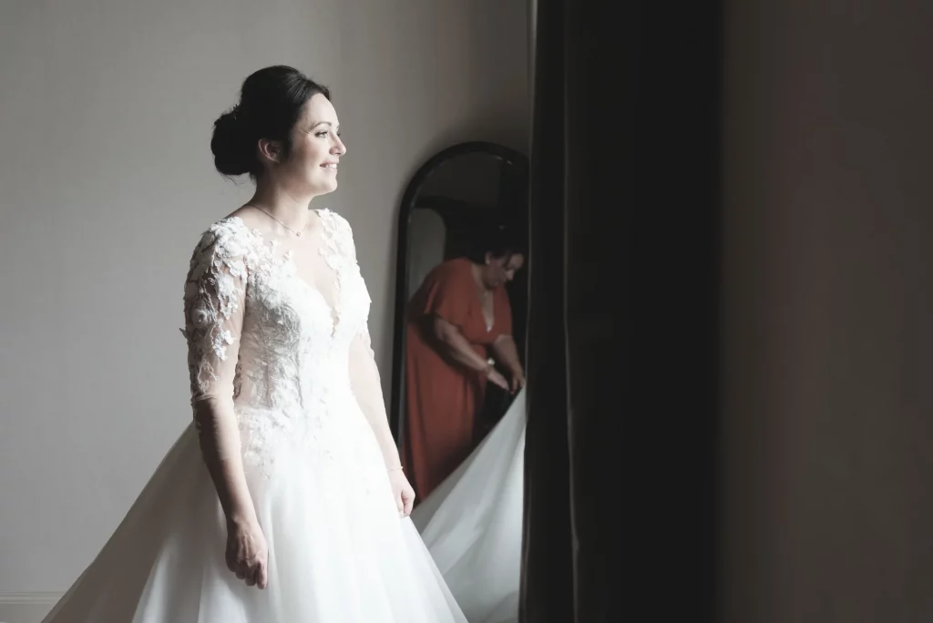 Mariée en robe de dentelle souriante à Saint-Hilaire-du-Harcouët, sa mère ajuste la robe dans le miroir.