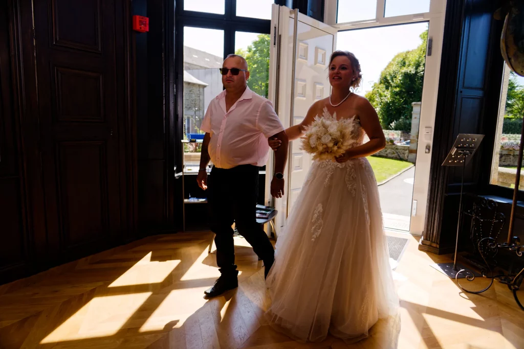 Mariée en robe dentelle et bouquet blanc entrant dans une salle en Normandie escortée par un homme.