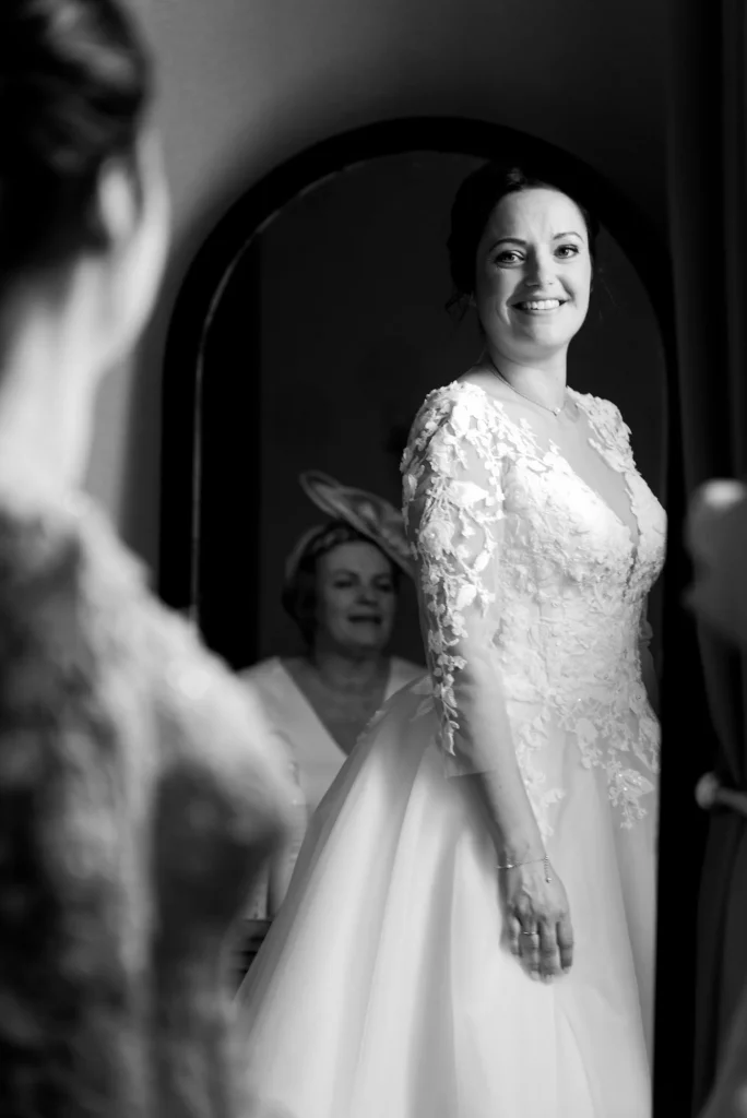 Mariée souriante devant un miroir, robe en dentelle, préparatifs de mariage à Saint-Hilaire-du-Harcouët.