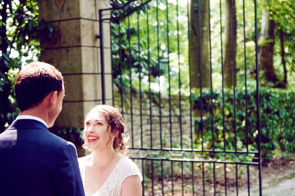 Mariée souriante regardant son époux devant une grille ancienne à Saint-Hilaire-du-Harcouët, Normandie.