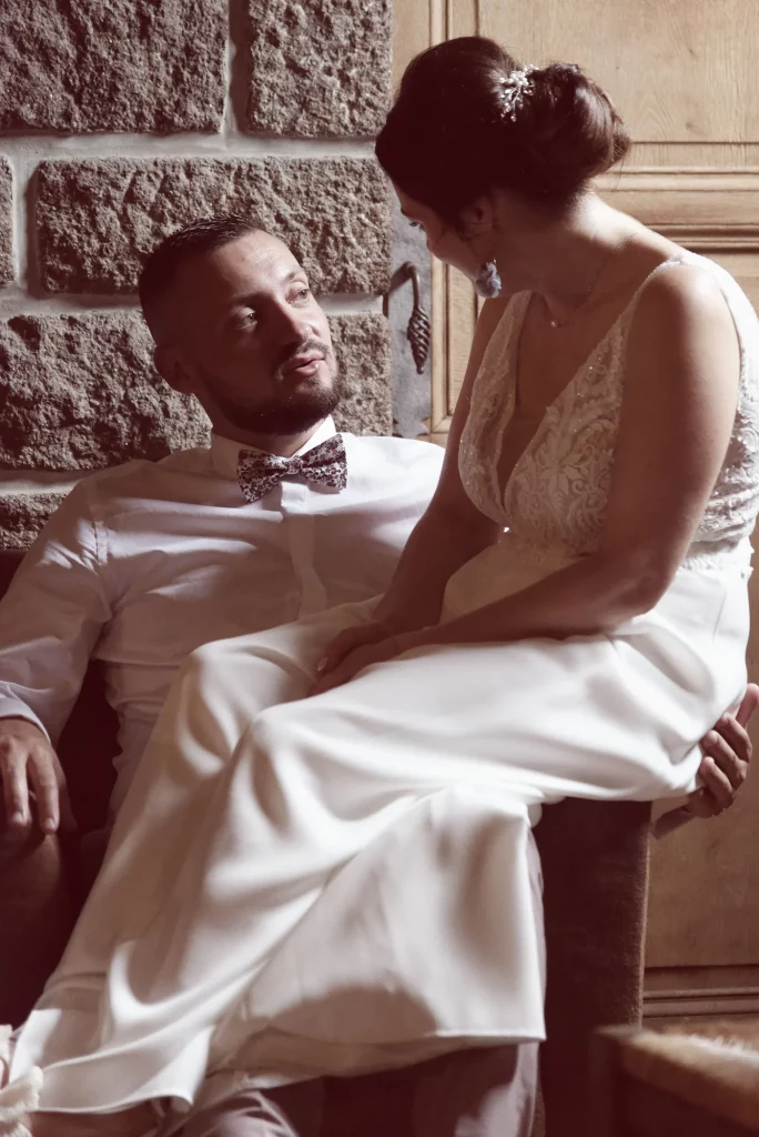 Mariée sur les genoux du marié devant un mur de pierre en Normandie, échange de regards complices.