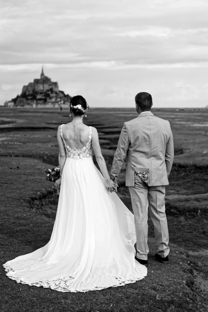 Mariés de dos main dans la main face au Mont Saint-Michel en Normandie. Photo noir et blanc.