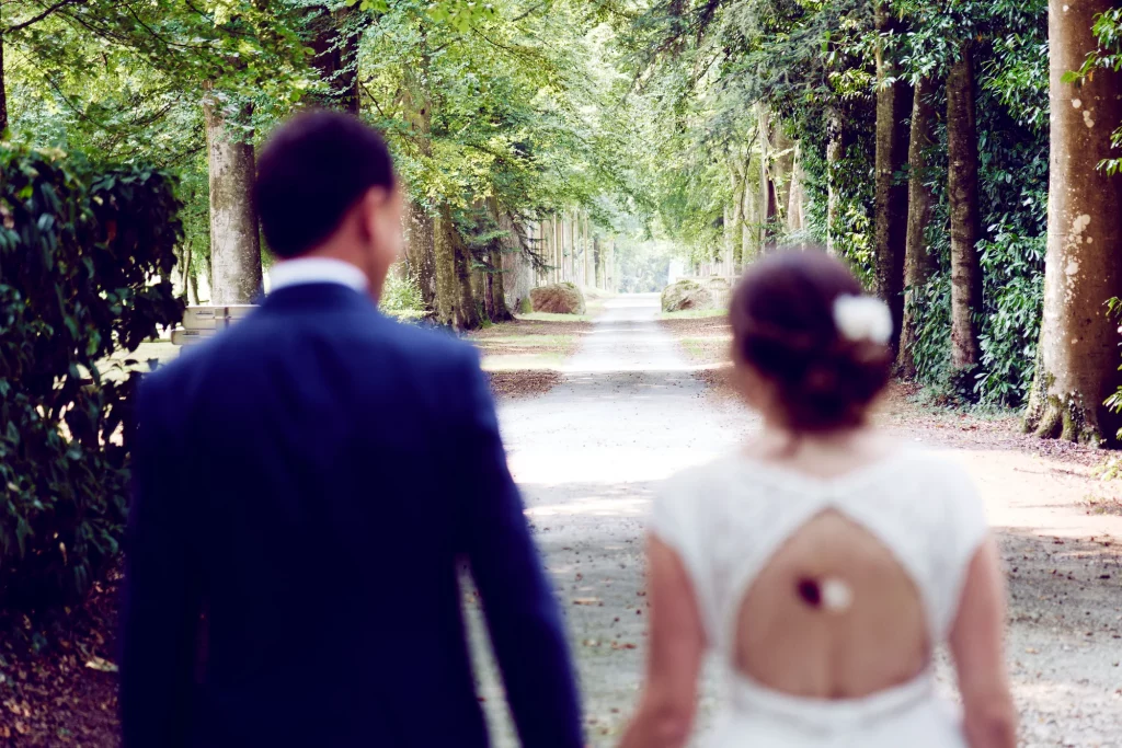 Mariés de dos marchant dans une allée d'arbres en Normandie, vers Saint-Hilaire-du-Harcouët.