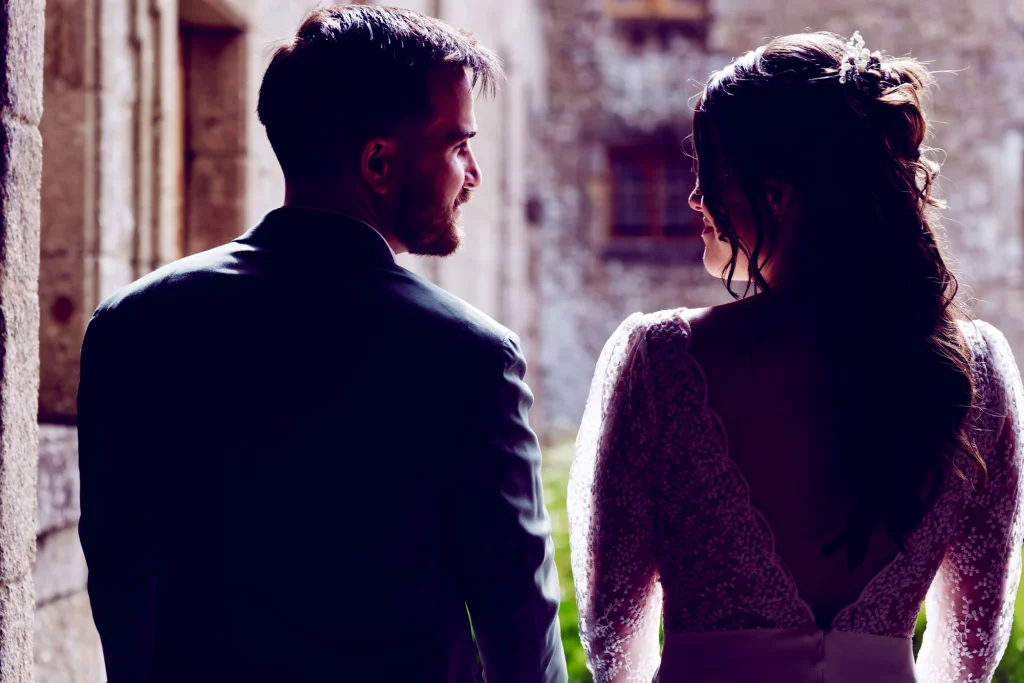 Mariés de dos se regardant devant un mur de pierre en Normandie, lumière cinématographique en contre-jour.