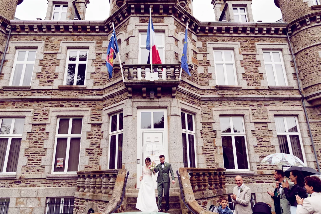 Mariés descendant l'escalier en pierre de la mairie de Saint-Hilaire-du-Harcouët en Normandie sous un parapluie.