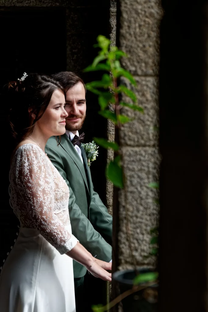 Mariés en Normandie, homme en costume vert et femme en robe dentelle près d'un mur en pierre à Avranches.