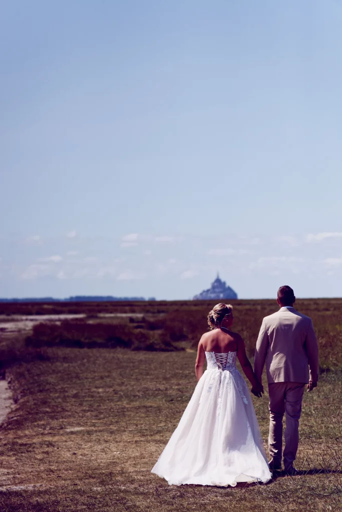 Mariés marchant main dans la main face au Mont Saint-Michel en Normandie, robe de mariée à lacets.