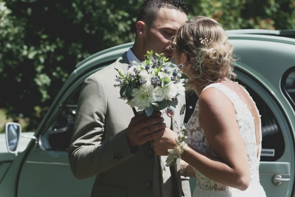 Mariés s'embrassant devant une 2CV à Saint-Hilaire-du-Harcouët avec un bouquet blanc et violet.