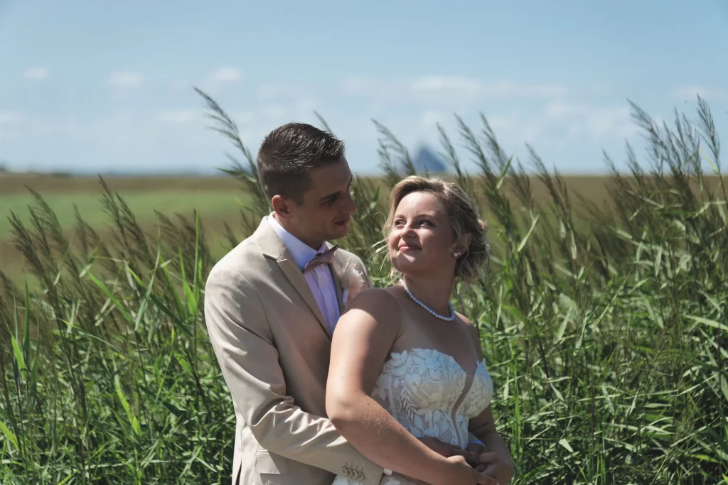 Mariés s'enlaçant dans les herbes hautes face au Mont Saint-Michel en Normandie, robe dentelle et costume beige.