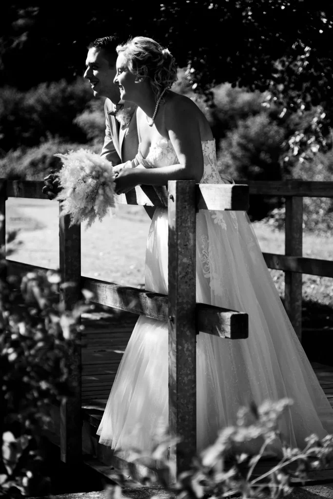 Mariés souriants sur un pont en bois à Saint-Hilaire-du-Harcouët en Normandie, noir et blanc contrasté.