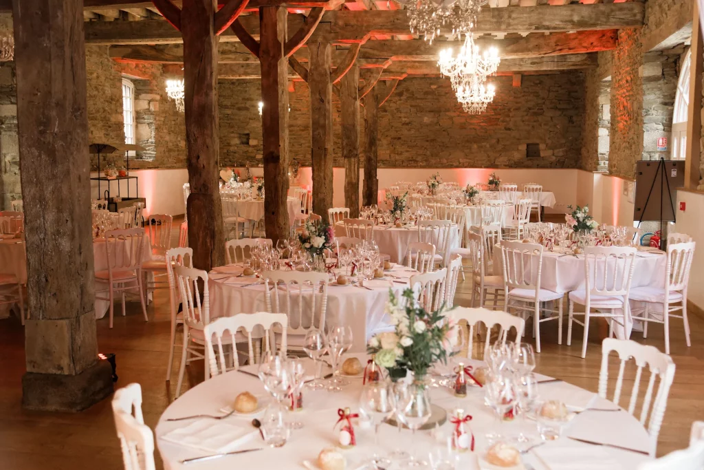 Salle de réception mariage Normandie avec pierres apparentes, tables blanches et lustres en cristal.