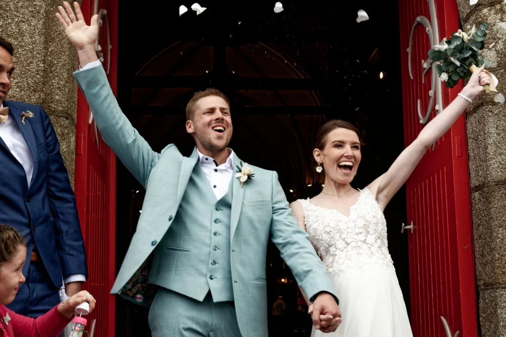 Sortie d'église en Normandie : mariés joyeux levant les bras sous des pétales devant une grande porte rouge.